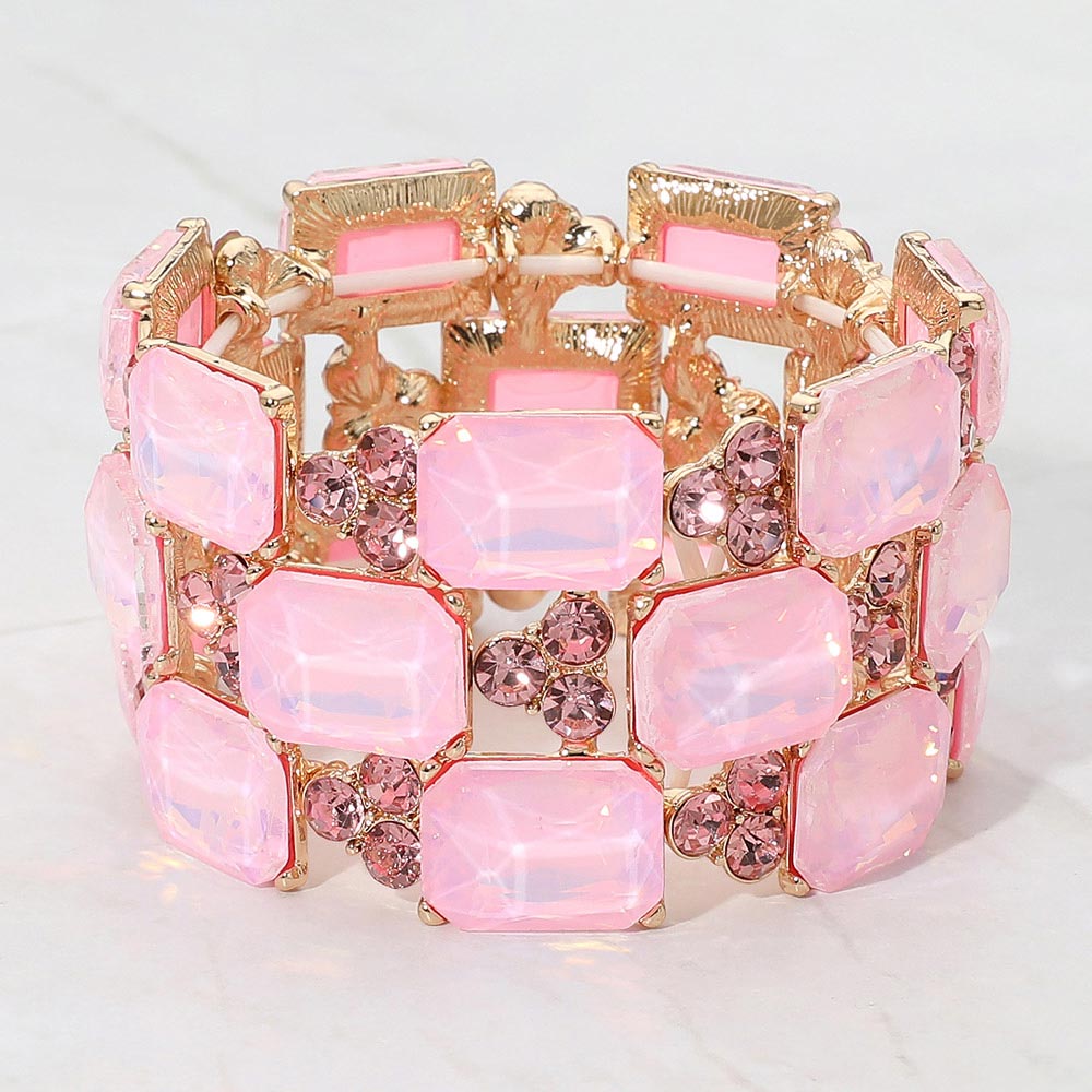 A gold-tone stretch bracelet featuring rectangular pink opal-like crystals and round darker pink faceted rhinestones, displayed on a light marble background.