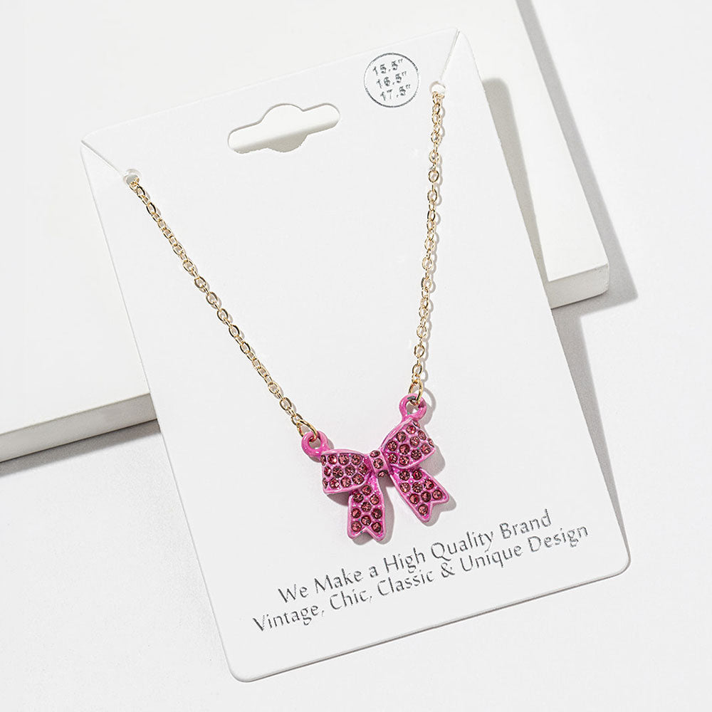 Bow pendant necklace with pink crystals in a pink setting on a gold-tone chain, measuring 15.5 inches with a 2-inch extender. The bow measures 0.75 inch wide by 0.75 inch long, with two rounded loops, a center knot, and two ribbon tails covered with small round crystals, displayed on a white jewelry card.