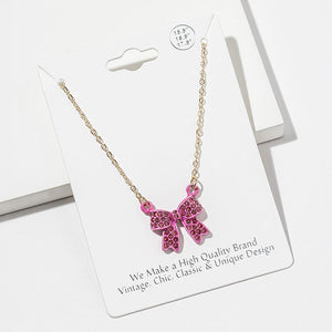 Bow pendant necklace with pink crystals in a pink setting on a gold-tone chain, measuring 15.5 inches with a 2-inch extender. The bow measures 0.75 inch wide by 0.75 inch long, with two rounded loops, a center knot, and two ribbon tails covered with small round crystals, displayed on a white jewelry card.