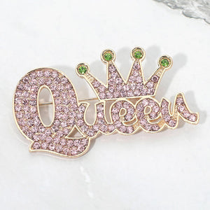 Pink and green crystal “Queen” brooch on gold-tone metal, featuring connected script lettering with a large looping capital Q and a five-point crown above the smaller letters. Measures approximately 2.75 inches wide by 1.5 inches tall, shown here on white marble. 
