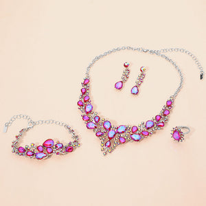 Fuchsia with reflective purple crystal 4-piece jewelry set on silver with statement necklace, matching bracelet, drop earrings and adjustable ring arranged on a light beige background.