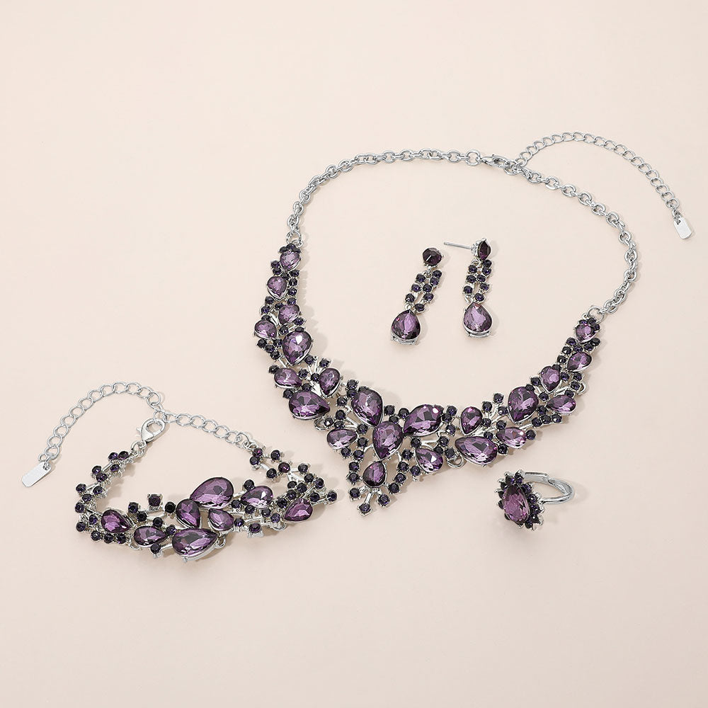 Purple crystal 4-piece jewelry set on silver with statement necklace, matching bracelet, drop earrings and adjustable ring arranged on a light beige background.