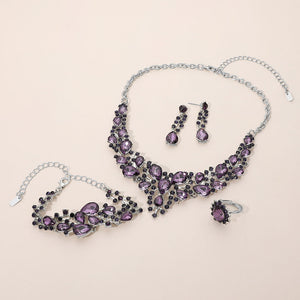 Purple crystal 4-piece jewelry set on silver with statement necklace, matching bracelet, drop earrings and adjustable ring arranged on a light beige background.