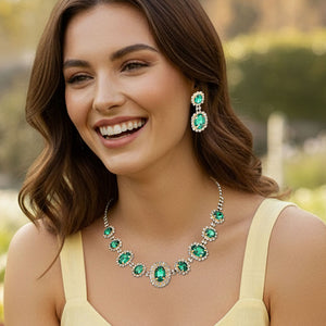 Jewelry set with emerald green gemstones surrounded by a halo of rhinestones on gold backing shown on a pretty lady outside on a blurred background.