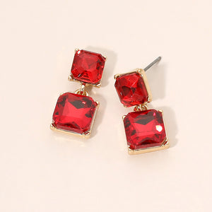 Red crystal double-square drop earrings on gold-tone metal, each featuring a smaller faceted square stone connected to a larger square stone below. Each earring measures approximately 0.4 inches wide by 0.9 inches long. Displayed on a pale peach surface.