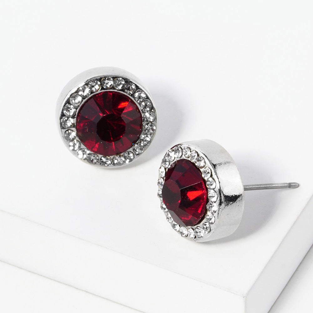 A pair of silver stud earrings with an oval red faceted center stone surrounded by a halo of small clear rhinestones, displayed on a clean white surface.