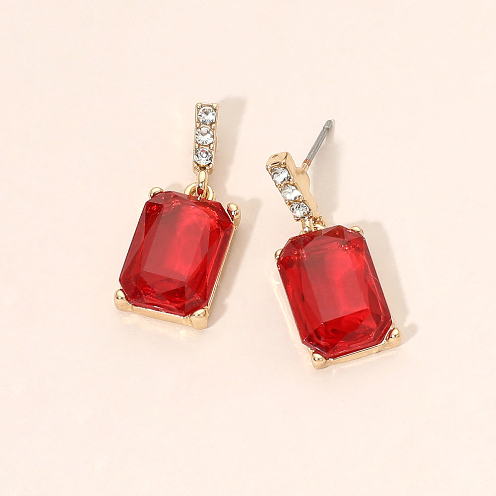 Gold-tone dangle earrings featuring large rectangular red crystals with a short row of small clear crystals above each stone, displayed on a light beige background.