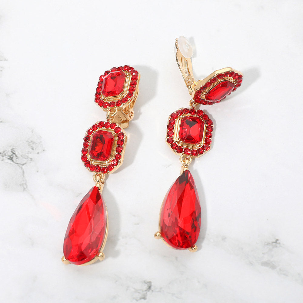 Red crystal square and teardrop clip-on earrings on gold-tone metal, displayed against a white marble background.