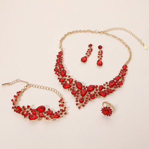 Red crystal 4-piece jewelry set on gold with statement necklace, matching bracelet, drop earrings and adjustable ring arranged on a light beige background.