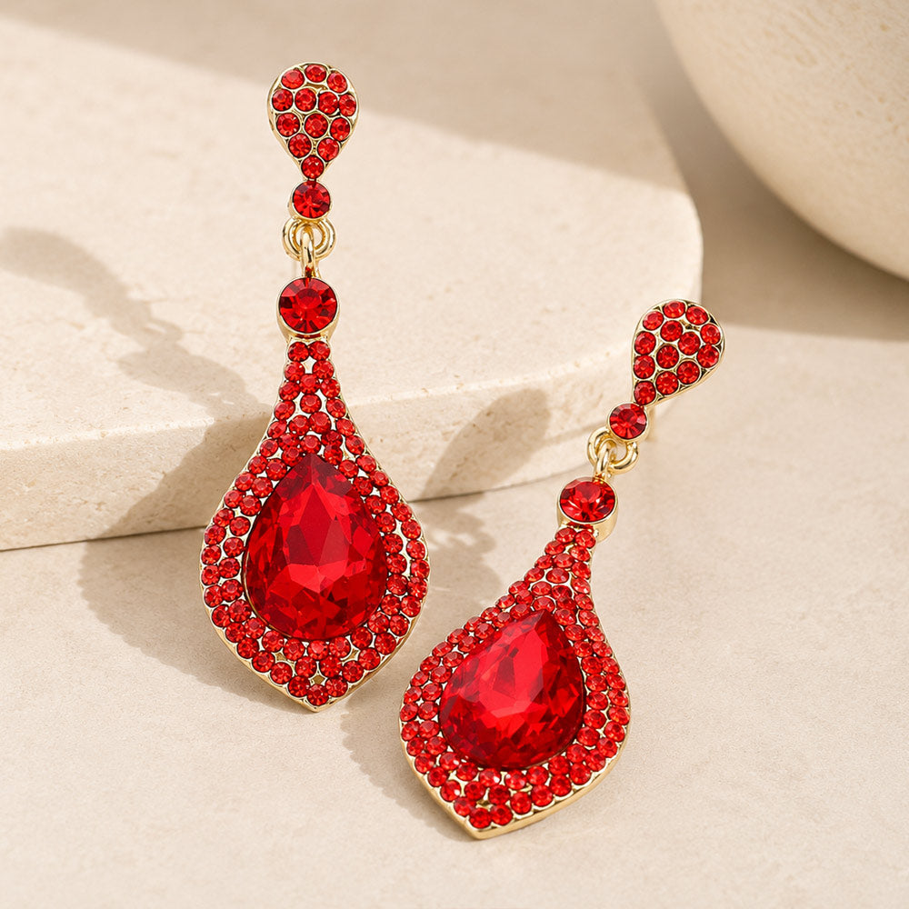 A pair of gold drop earrings adorned with red crystals, featuring a large teardrop crystal surrounded by smaller red crystals, displayed on a neutral beige background.