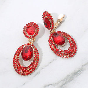 Red oval double-halo crystal clip-on earrings on gold on a white marble background — 2.1" statement drops for evening or pageant glam.