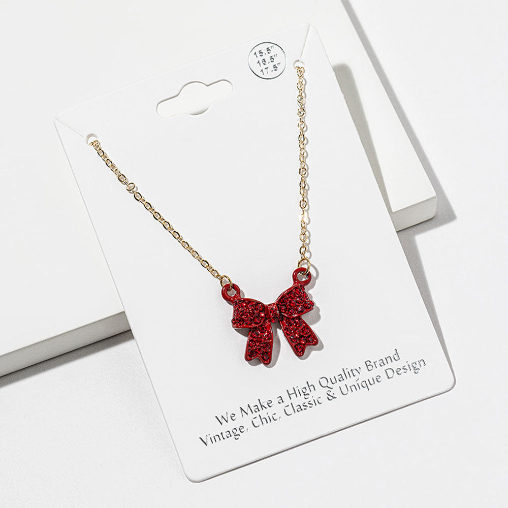 Bow pendant necklace with red crystals in a red setting on a gold-tone chain, measuring 15.5 inches with a 2-inch extender. The 0.75-inch-wide and long bow has two rounded loops, a center knot, and two ribbon tails covered with small round crystals, displayed on a white jewelry card.