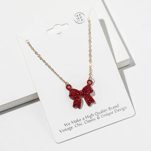 Bow pendant necklace with red crystals in a red setting on a gold-tone chain, measuring 15.5 inches with a 2-inch extender. The 0.75-inch-wide and long bow has two rounded loops, a center knot, and two ribbon tails covered with small round crystals, displayed on a white jewelry card.