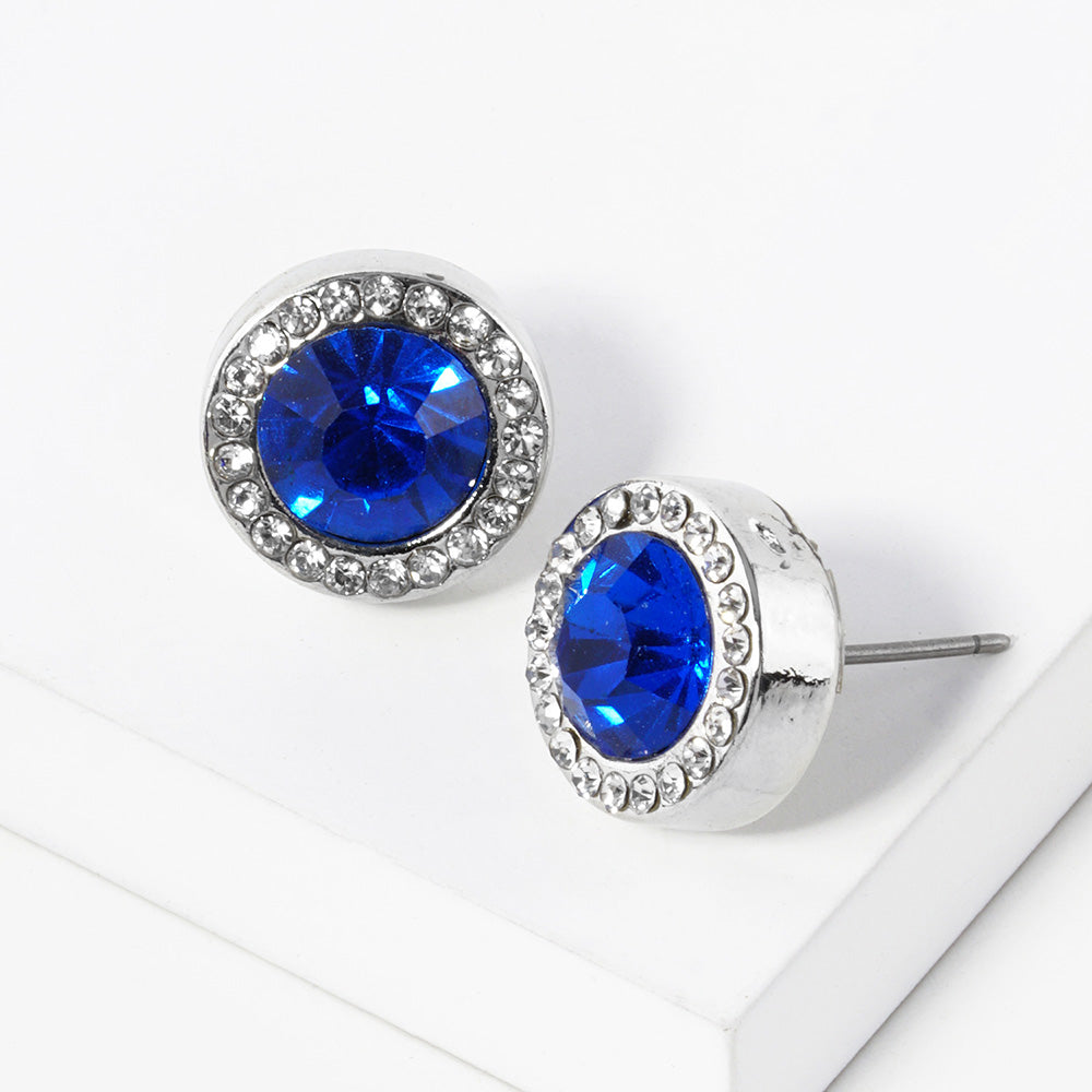 A pair of silver stud earrings with an oval royal blue faceted center stone surrounded by a halo of small clear rhinestones, displayed on a clean white surface.