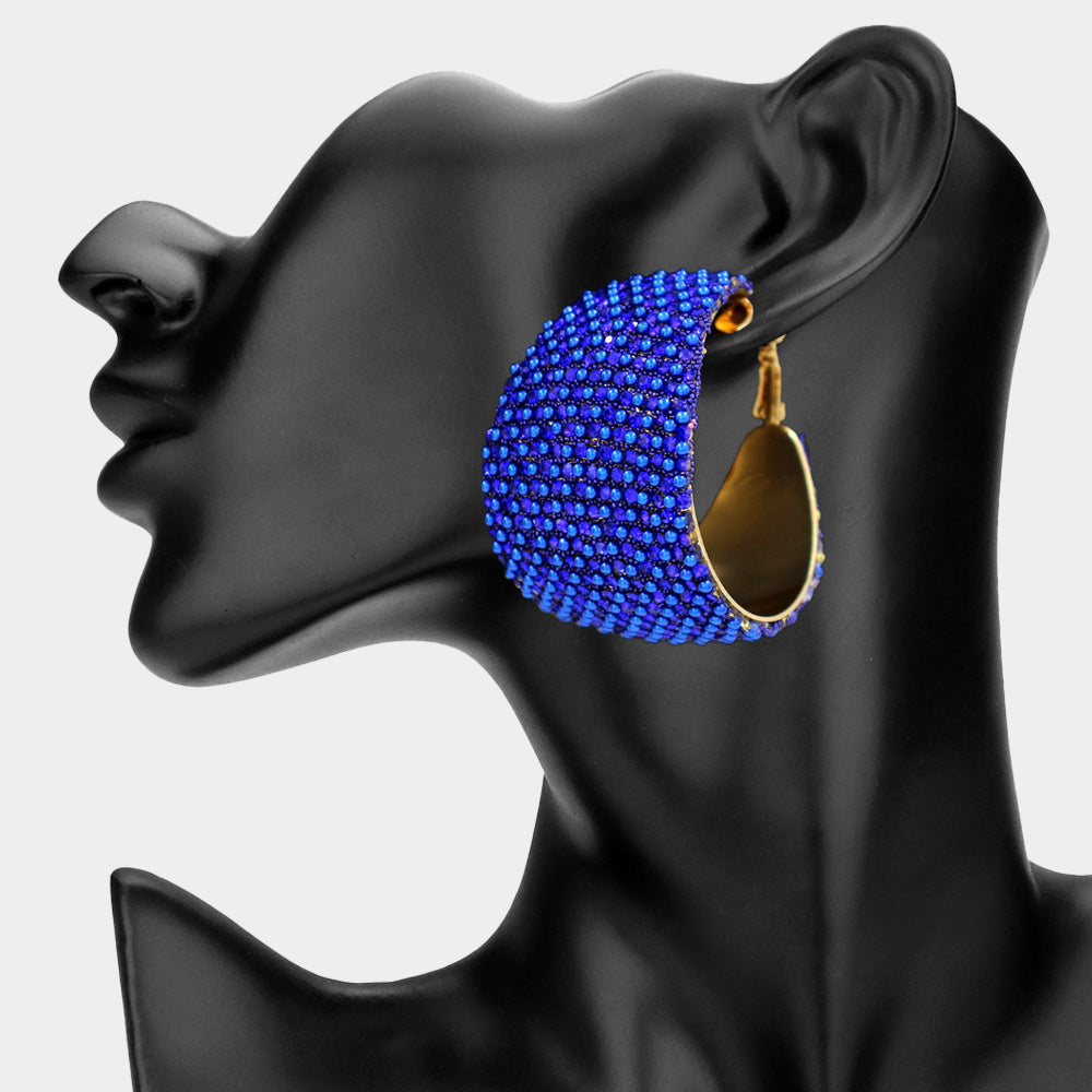 Sapphire royal blue wide crystal hoop earrings on gold-tone metal with dense faceted rhinestone coverage and omega backs, displayed on a black mannequin profile.