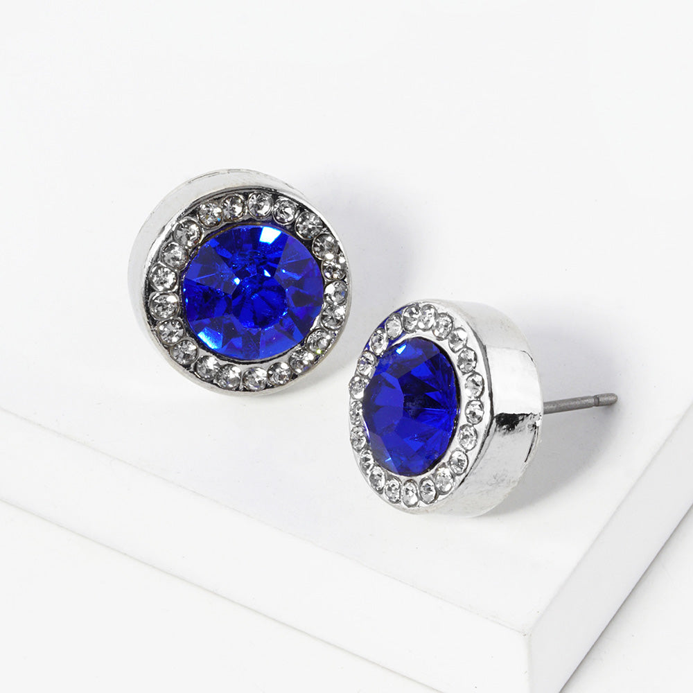 A pair of silver stud earrings with an oval sapphire blue faceted center stone surrounded by a halo of small clear rhinestones, displayed on a clean white surface.