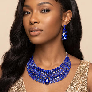 Sapphire royal blue crystal statement necklace set on silver-tone metal, featuring a broad, layered bib of marquise and round stones with a large teardrop centerpiece and matching teardrop earrings. Necklace measures 17 inches plus a 2-inch extender, with 2.25-inch decor; earrings are 2.2 inches long. Worn by a woman in a gold sequined outfit against a warm beige background.