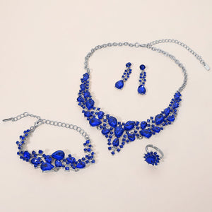 Sapphire royal blue crystal 4-piece jewelry set on silver with statement necklace, matching bracelet, drop earrings and adjustable ring arranged on a light beige background.