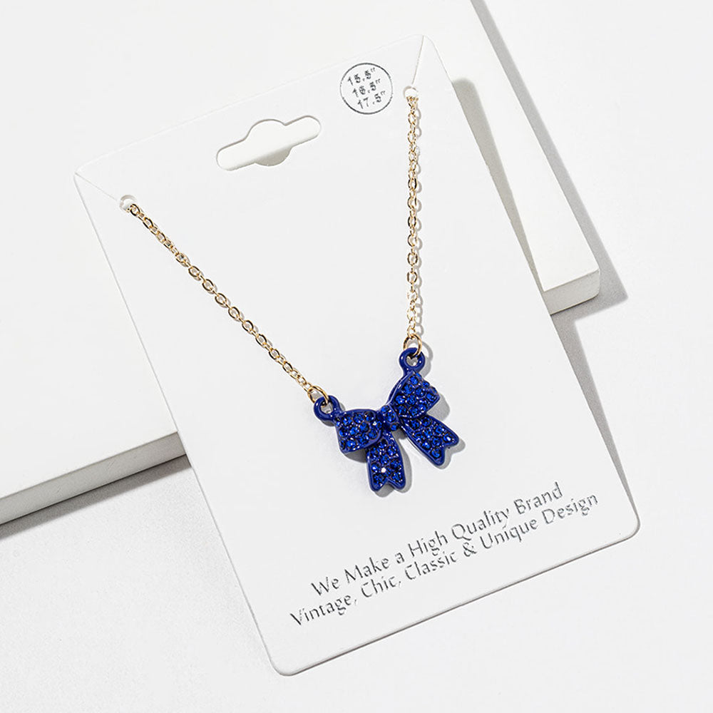 Bow pendant necklace with sapphire royal blue crystals in a blue setting on a gold-tone chain, measuring 15.5 inches with a 2-inch extender. The bow measures 0.75 inch wide by 0.75 inch long, with two rounded loops, a center knot, and two ribbon tails covered with small round crystals, displayed on a white jewelry card.