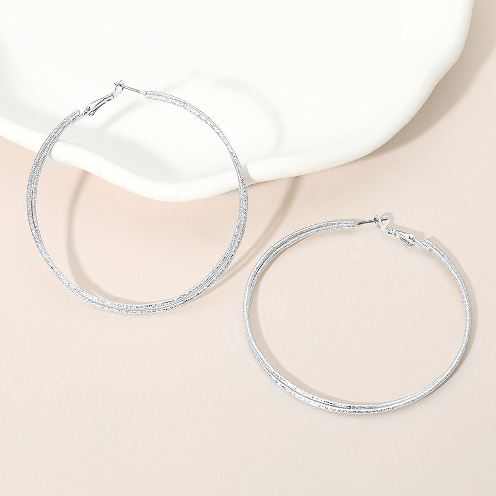 Silver textured double hoop earrings with a 2.75-inch diameter and pin catch clasp, displayed on a light pink background.