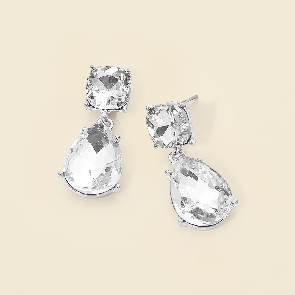 Clear crystal drop earrings on silver with a round top stone and large teardrop crystal, displayed on a light cream background.