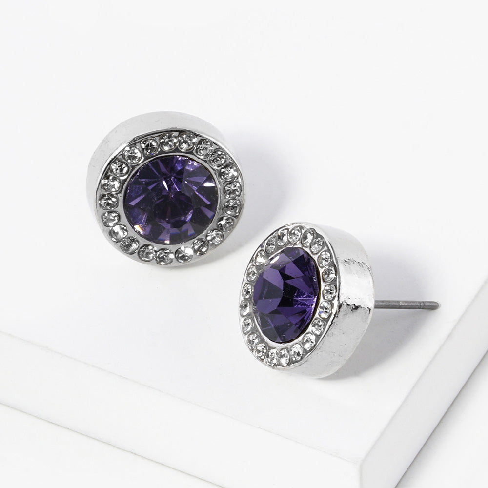 A pair of silver stud earrings with an oval purple faceted center stone surrounded by a halo of small clear rhinestones, displayed on a clean white surface.