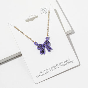 Bow pendant necklace with purple crystals in a purple setting on a gold-tone chain, measuring 15.5 inches with a 2-inch extender. The bow measures 0.75 inch wide by 0.75 inch long, with two rounded loops, a center knot, and two ribbon tails covered with small round crystals, displayed on a white jewelry card.