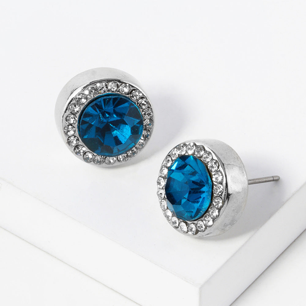 A pair of silver stud earrings with an oval teal faceted center stone surrounded by a halo of small clear rhinestones, displayed on a clean white surface.