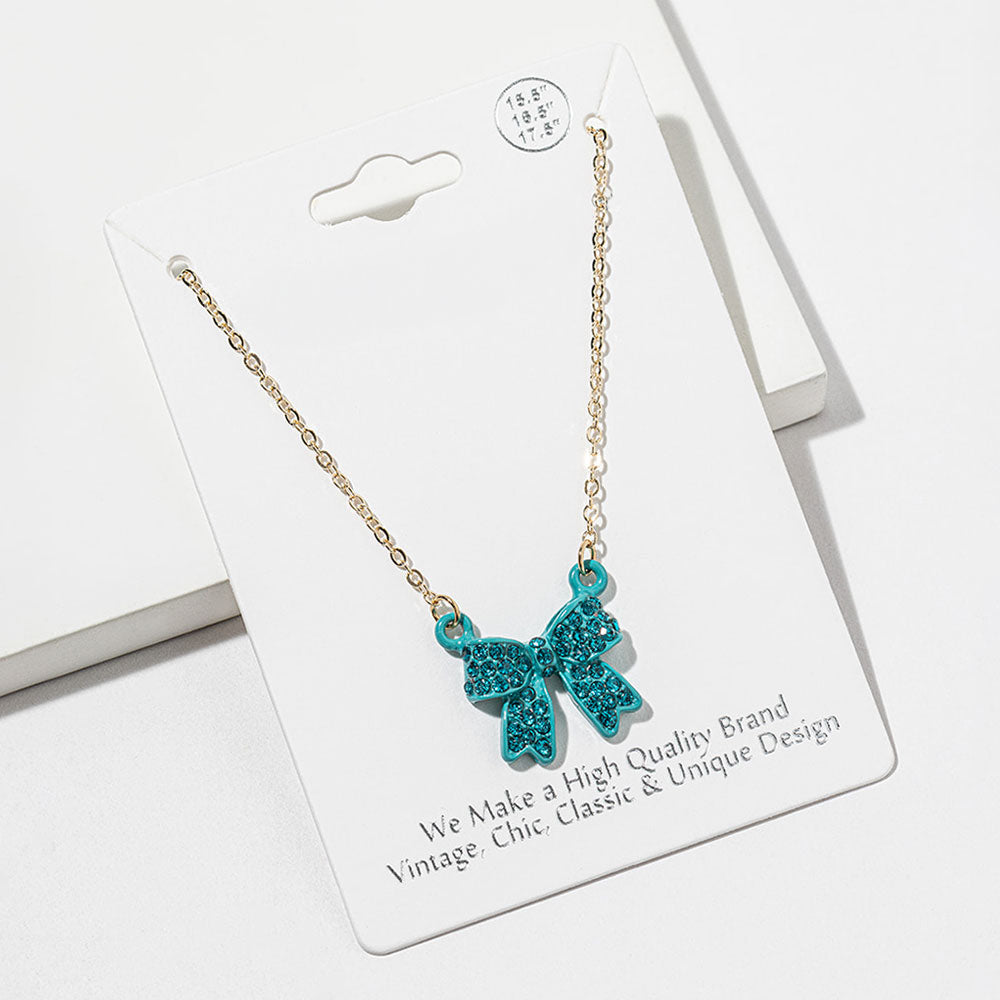 Bow pendant necklace with teal crystals in a teal setting on a gold-tone chain, measuring 15.5 inches with a 2-inch extender. The bow measures 0.75 inch wide by 0.75 inch long, with two rounded loops, a center knot, and two ribbon tails covered with small round crystals, displayed on a white jewelry card.