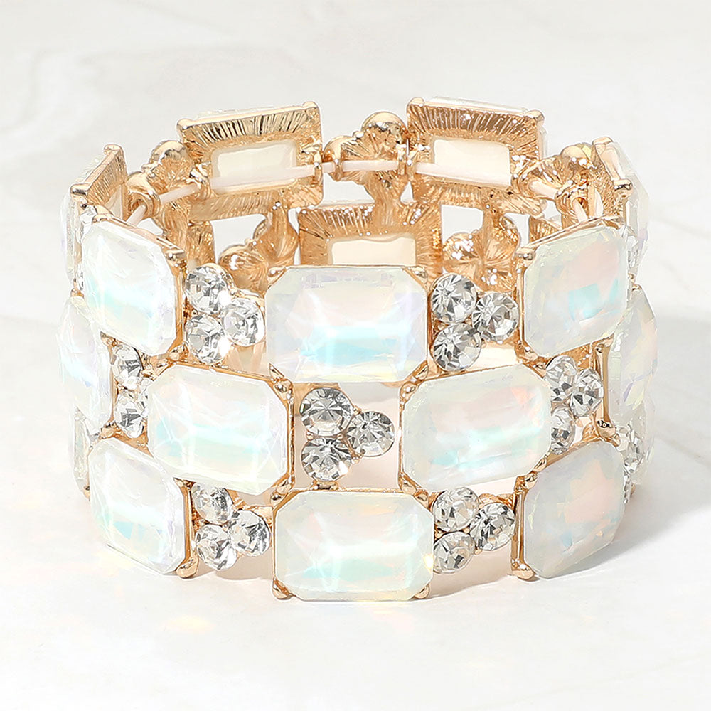 A gold-tone stretch bracelet featuring rectangular white opal-like crystals and round clear faceted rhinestones, displayed on a light marble background.