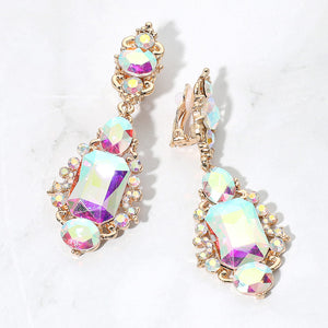 Iridescent AB crystals reflecting pink, yellow, and pale blue in vintage-inspired clip-on earrings on gold-tone settings, measuring 1 inch wide by 2.7 inches long. Decorative oval crystal tops connect to drops with large rectangular center stones, smaller round and oval stones, and curved metal details. One clip-on fastening is visible, displayed on a white textured surface.