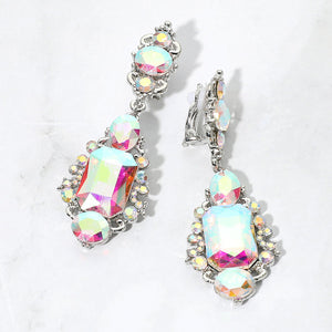 Iridescent AB crystals reflecting pink, yellow, and pale blue in vintage-inspired clip-on earrings on silver-tone settings, measuring 1 inch wide by 2.7 inches long. Decorative oval crystal tops connect to drops with large rectangular center stones, smaller round and oval stones, and curved metal details. One clip-on fastening is visible, displayed on a white textured surface.