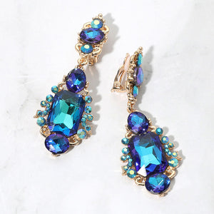 Blue AB crystals with teal and purple reflections in vintage-inspired clip-on earrings on gold-tone settings, measuring 1 inch wide by 2.7 inches long. Decorative oval crystal tops connect to drops with large rectangular center stones, smaller round and oval stones, and curved metal details. One clip-on fastening is visible, displayed on a white textured surface.