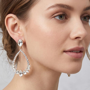 Clear crystal open teardrop earrings on gold-tone settings, measuring 1.25 inches wide by 2.6 inches long. A teardrop top connects to an open crystal-lined drop with larger faceted stones along the bottom. Shown in the clear color on a model to illustrate the style’s size and placement beside the face, against a light gray background.