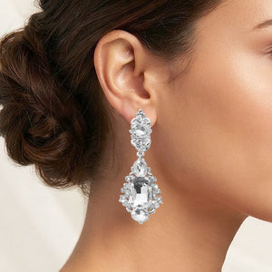 Clear crystal vintage-inspired clip-on earrings on silver-tone settings, measuring 1 inch wide by 2.7 inches long. An ornate oval crystal cluster connects to a drop with a large rectangular center stone, a teardrop above, and a round stone below, shown on a model with her hair in a low bun against a cream background.