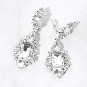 Pair of clear crystal vintage-inspired clip-on earrings on silver-tone settings, measuring 1 inch wide by 2.7 inches long. Oval crystal tops connect to rectangular center stones framed by small round crystals and curved metal details, with teardrops above and round stones below. One clip-on fastening is visible, displayed on a white textured surface.