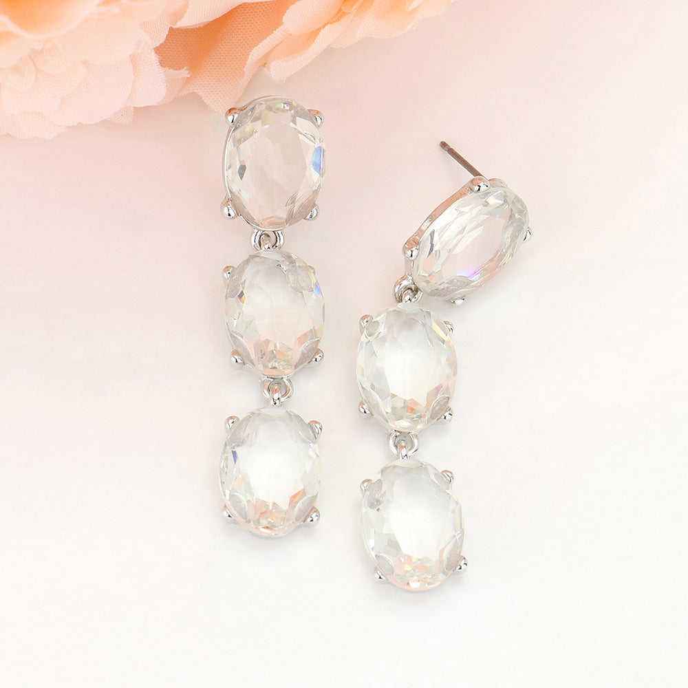 Triple oval drop earrings with three linked, faceted clear glass stones in silver-tone settings and post backs, approximately 0.4 inches wide and 2 inches long, displayed against a pale background with peach flowers.