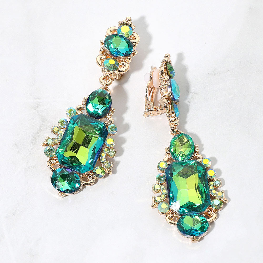 Green AB crystals with teal and yellow-green reflections in vintage-inspired clip-on earrings on gold-tone settings, measuring 1 inch wide by 2.7 inches long. Decorative oval crystal tops connect to drops with large rectangular center stones, smaller round and oval stones, and curved metal details. One clip-on fastening is visible, displayed on a white textured surface.