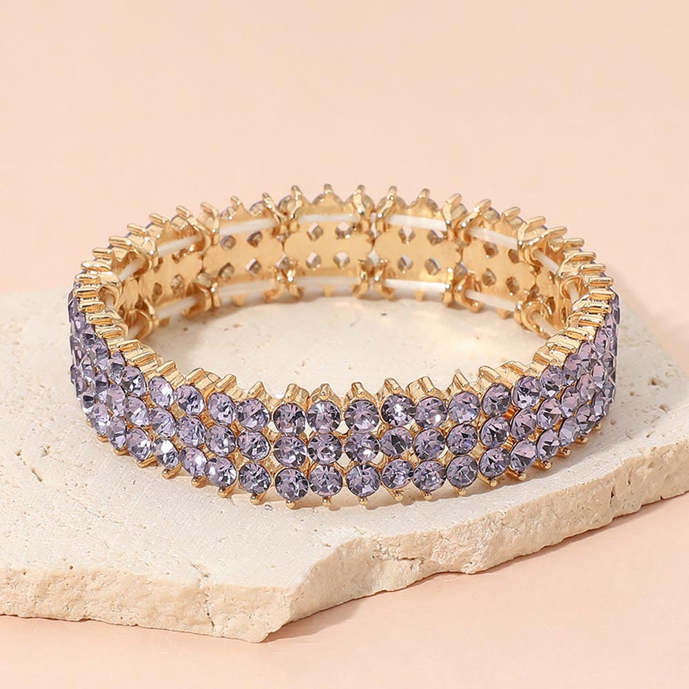 Lavender crystal stretch bracelet on gold-tone metal, approximately 0.5 inches high, with three rows of round faceted pale purple stones. Displayed on a textured light beige stone slab against a soft peach-pink background.