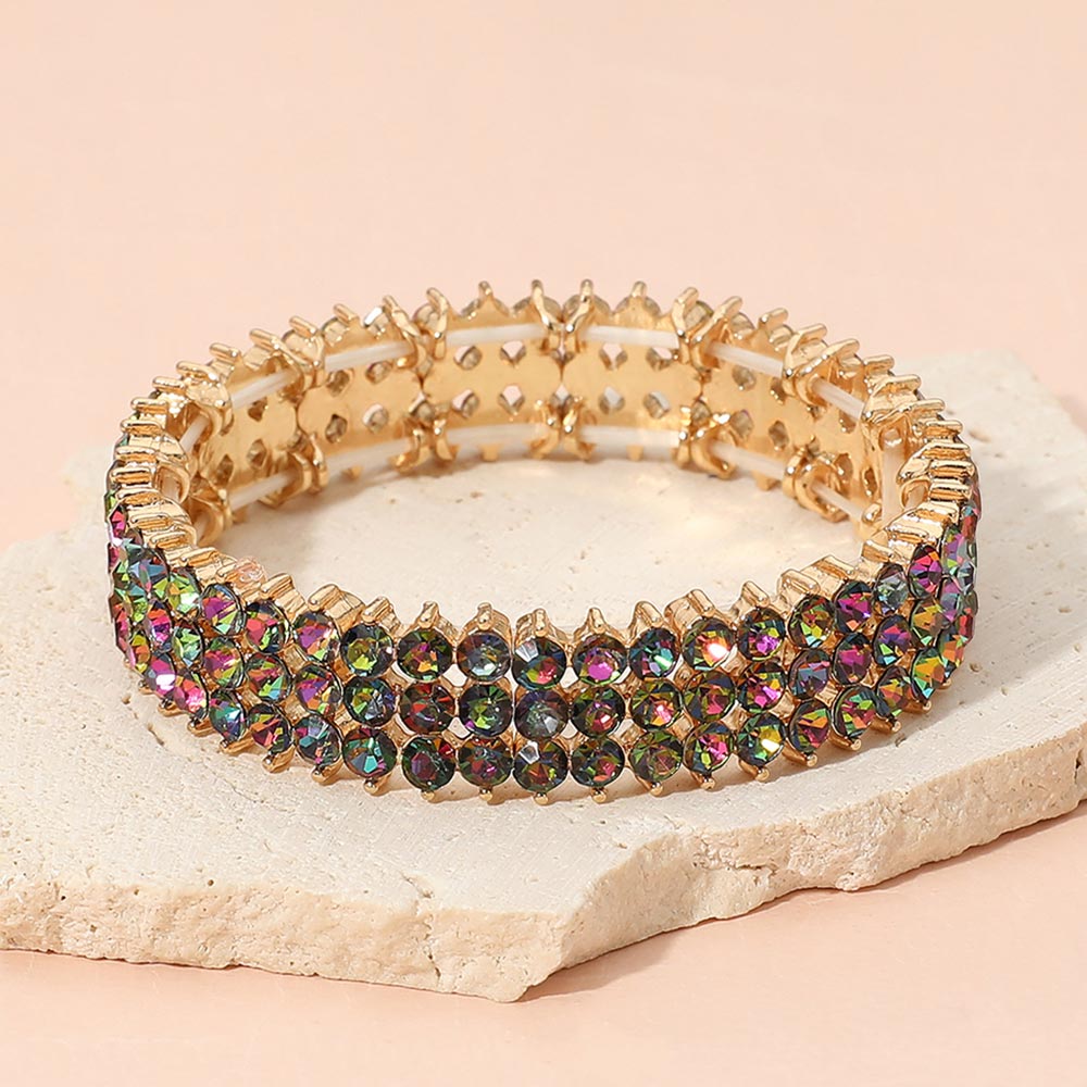 Multi-color crystal stretch bracelet on gold-tone metal, approximately 0.5 inches high, with three rows of round faceted stones reflecting deep pink, green, gold, and other colors. Displayed on a textured light beige stone slab against a soft peach-pink background.