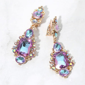 Multi-color crystals reflecting purple, pink, and blue in vintage-inspired clip-on earrings on gold-tone settings, measuring 1 inch wide by 2.7 inches long. Decorative oval crystal tops connect to drops with large rectangular center stones, smaller round and oval stones, and curved metal details. One clip-on fastening is visible, displayed on a white textured surface.