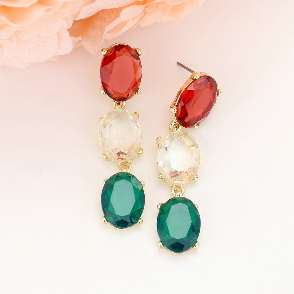 Triple oval drop earrings with three linked, faceted red, clear, and green glass stones in gold-tone settings and post backs, approximately 0.4 inches wide and 2 inches long, displayed against a pale background with peach flowers.
