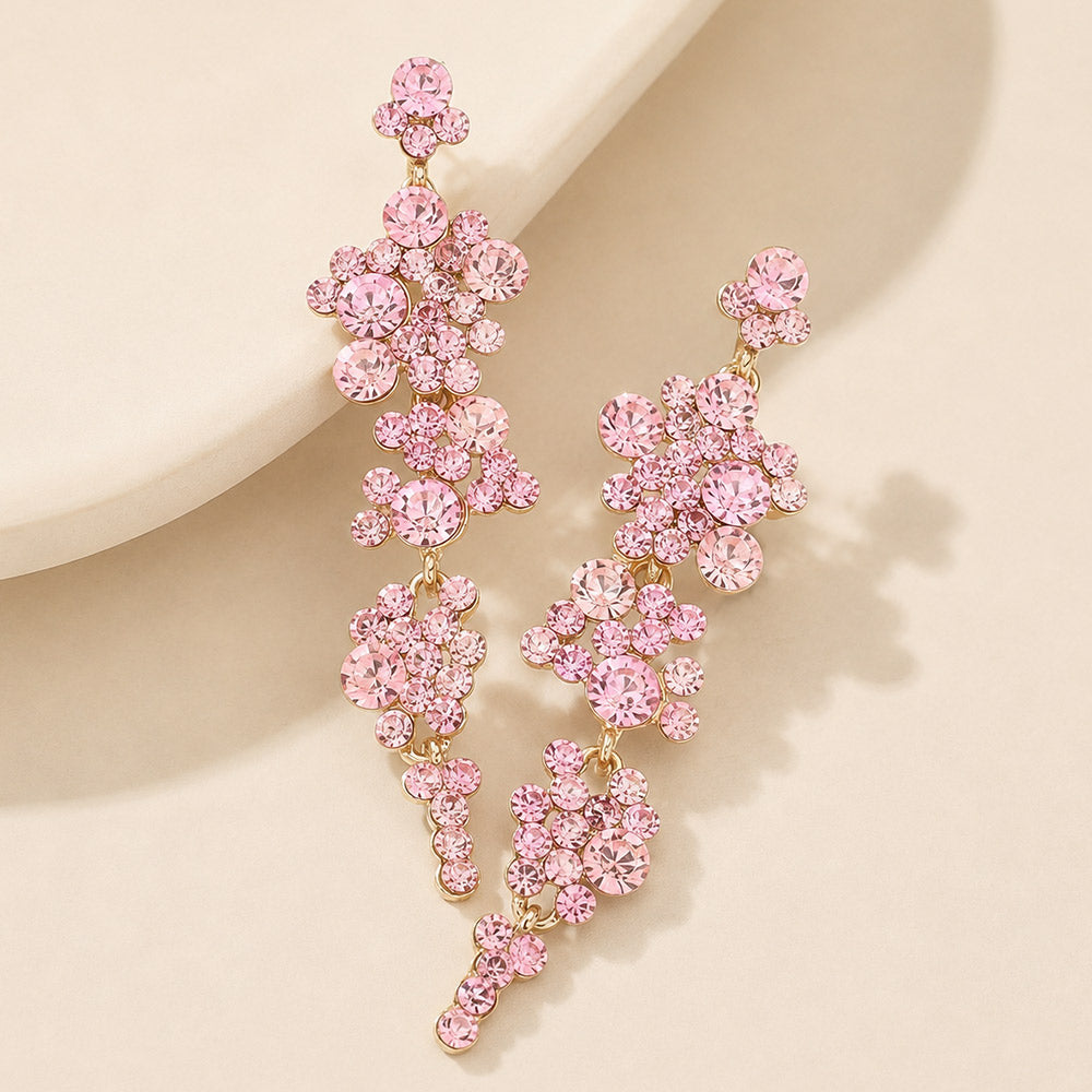 Pink crystal long dangle earrings on gold-tone settings, with linked clusters of round faceted stones tapering to small crystal tips, arranged on a beige display with soft shadows.