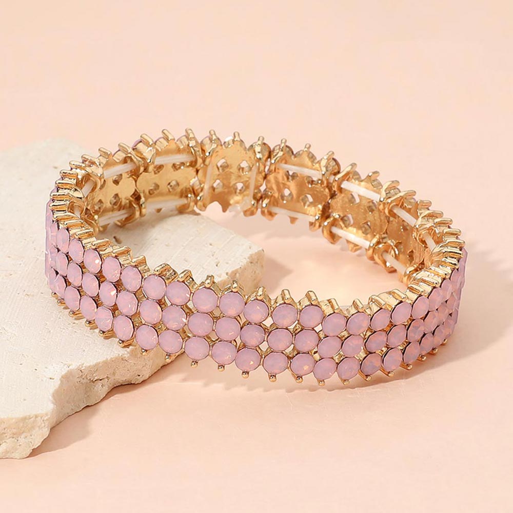 Pink opal crystal stretch bracelet on gold-tone metal, approximately 0.5 inches high, with three rows of round pale pink stones. Displayed on a textured light beige stone slab against a soft peach-pink background.